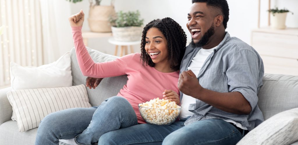Couple Leisure. Happy Black Spouses Watching Tv And Cheering Sports, Eating Popcorn And Relaxing On Couch In Living Room