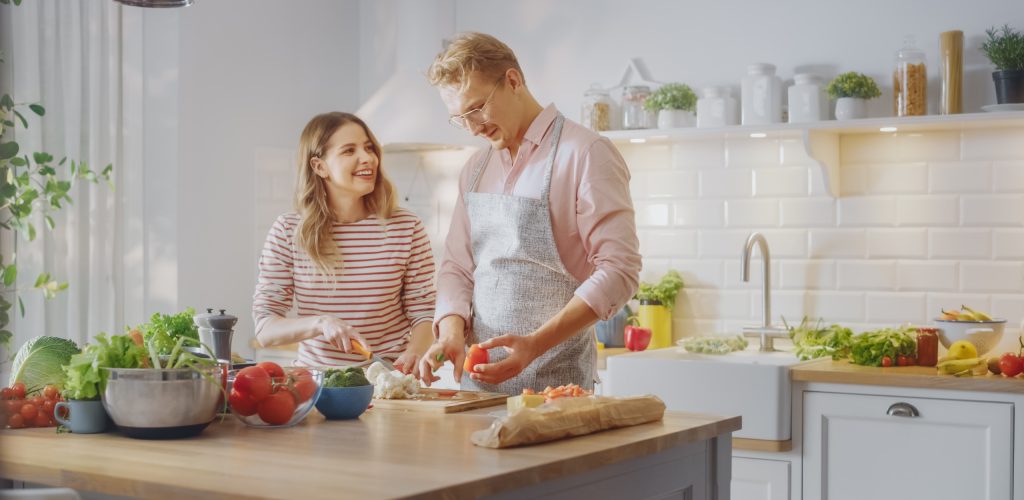 Handsome Young Man in Glasses Wearing Apron and Beautiful Girl are Preparing a Salad in the Kitchen. Happy Couple are Casually Talking. Natural Clean Diet and Healthy Way of Life Concept.